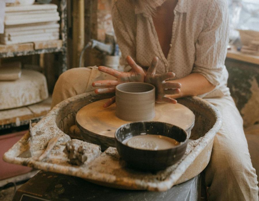 A person shapes clay on a pottery wheel in a cozy studio filled with shelves and pottery tools.