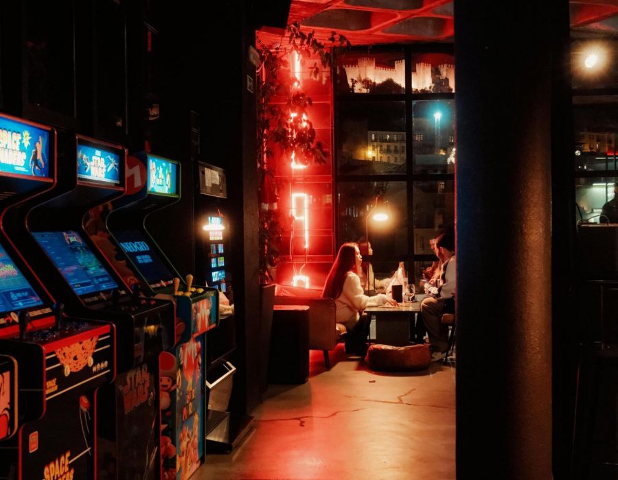 Dimly lit arcade with glowing game machines and people sitting near a window with red neon lights.