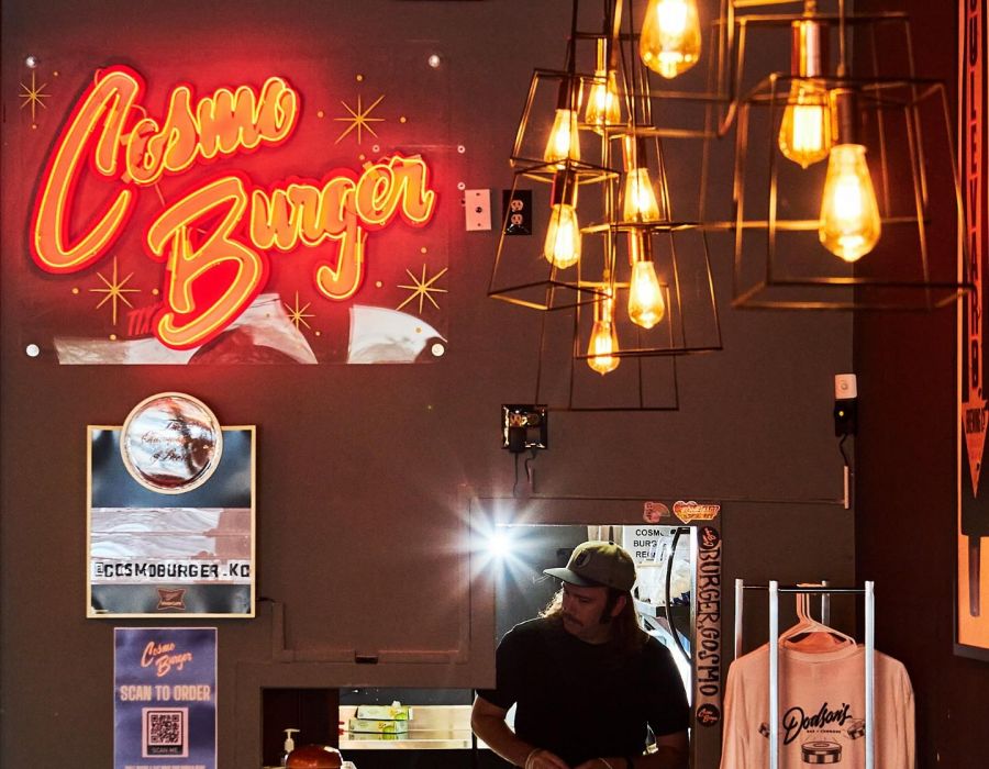 A person works at a counter under hanging lights, with a neon "Chelsea Burger" sign on the wall.